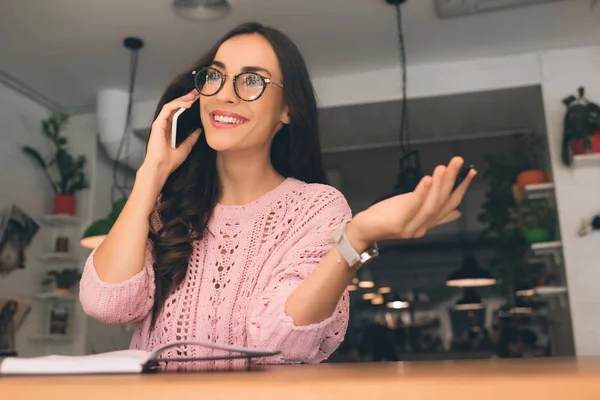Glückliche junge Frau mit Brille Frau spricht auf Smartphone mit Lehrbuch am Tisch im Café — Stockfoto