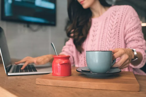Cropped image of female freelancer sitting with laptop and coffee cup at table in cafe — Stock Photo