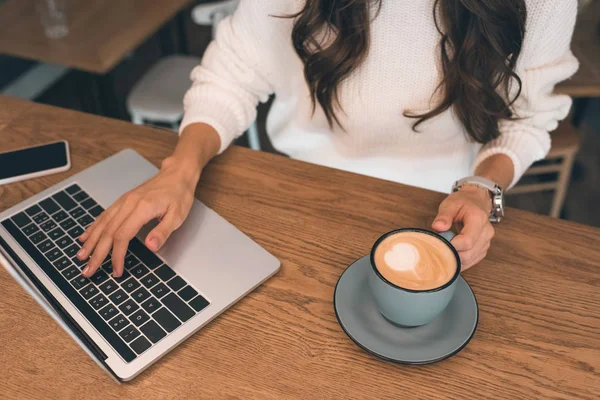 Vista parziale di una donna freelance che lavora su un computer portatile mentre siede a tavola con smartphone e tazza di caffè nel caffè — Foto stock