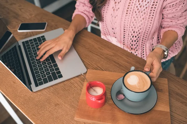 Imagen recortada del freelancer femenino que trabaja en el ordenador portátil mientras está sentado en la mesa con teléfono inteligente y taza de café en la cafetería - foto de stock