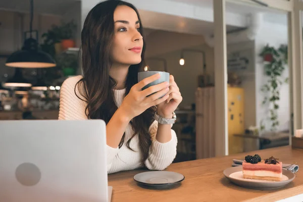 Attractive female freelancer sitting with laptop, cake and coffee cup at table in cafe — Stock Photo