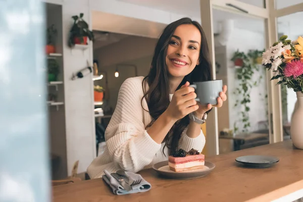 Heureuse jeune femme avec tasse de café assis à table avec gâteau au fromage dans le café — Photo de stock