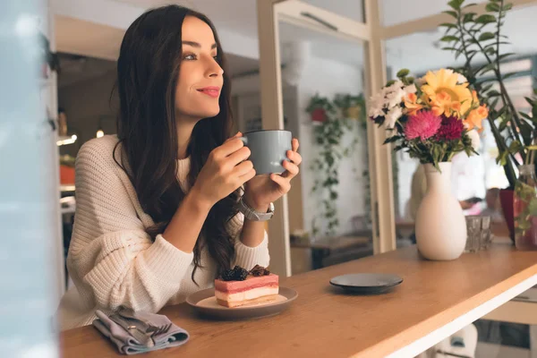 Mujer joven pensativa con taza de café sentado en la mesa con pastel de queso en la cafetería - foto de stock