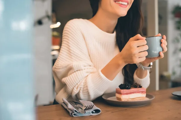 Vista parcial de la mujer sonriente con taza de café sentado en la mesa con pastel de queso en la cafetería - foto de stock