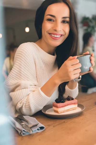 Sorridente giovane donna con tazza di caffè seduta a tavola con cheesecake nel caffè — Foto stock