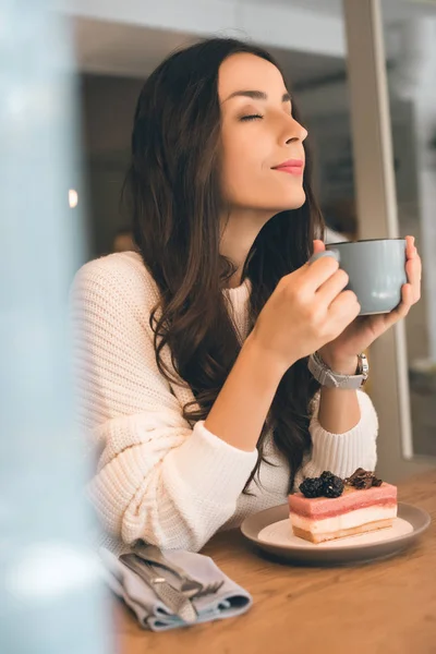 Atractiva joven con los ojos cerrados sosteniendo taza de café y sentado a la mesa con pastel de queso en la cafetería - foto de stock