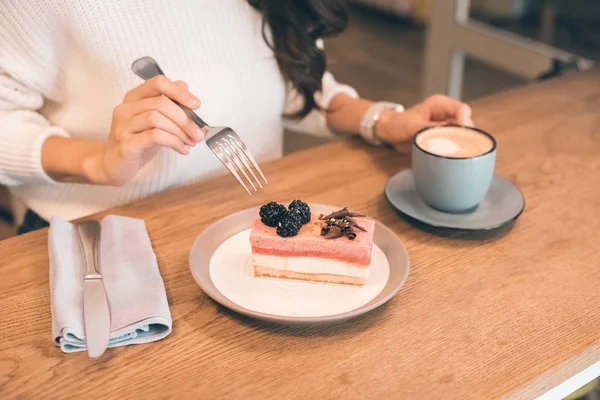 Immagine ritagliata di giovane donna con tazza di caffè mangiare cheesecake a tavola nel caffè — Foto stock