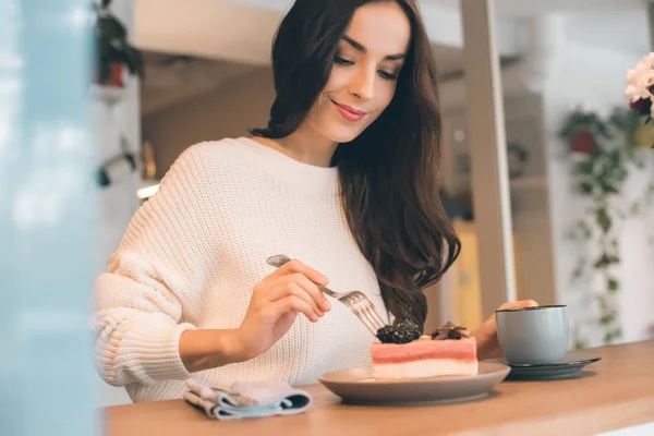 Selective focus of young woman with coffee cup eating cheesecake at table in cafe — Stock Photo