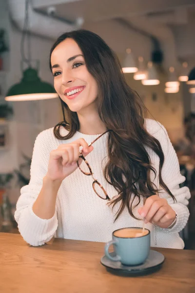Весела молода жінка тримає окуляри, сидячи за столом з чашкою кави в кафе — стокове фото