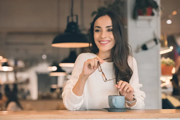 Улыбающаяся молодая женщина, держащая очки, сидя за столом с чашкой кофе в кафе — стоковое фото