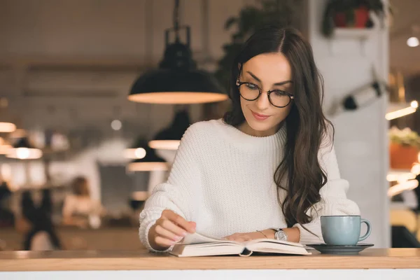 Attractive young woman in eyeglasses reading book at table with coffee cup in cafe — Stock Photo