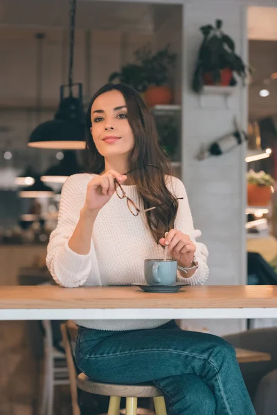 Bela jovem mulher olhando para longe e segurando óculos enquanto sentado à mesa com xícara de café no café — Fotografia de Stock