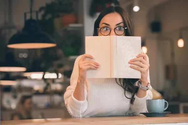 Шокована жінка в окулярах, що покриває обличчя за книгою за столом з чашкою кави в кафе — стокове фото