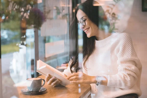 Laughing young woman reading book at table with coffee cup in cafe — Stock Photo