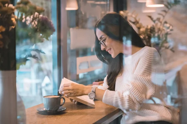 Smiling woman reading book at table with coffee cup in cafe — Stock Photo