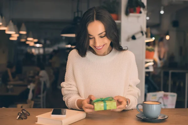 Приваблива щаслива жінка тримає подарункову коробку за столом з чашкою кави та смартфоном у кафе — стокове фото