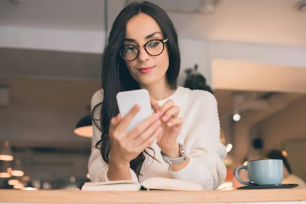 Красива молода жінка в окулярах використовує смартфон за столом з чашкою кави в кафе — стокове фото