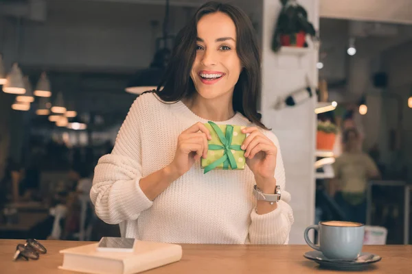 Mujer joven feliz mostrando caja de regalo en la mesa con teléfono inteligente y taza de café en la cafetería - foto de stock