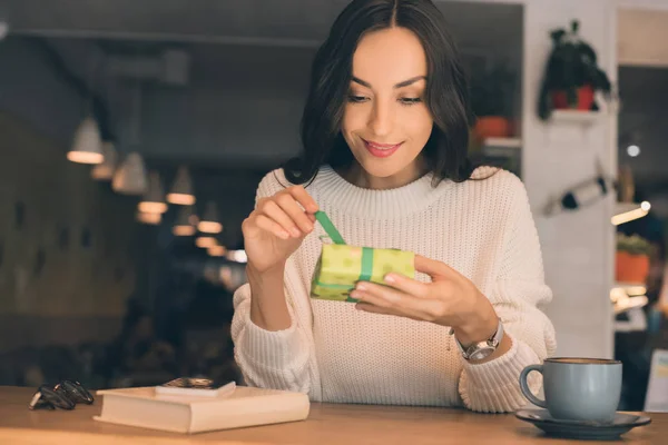 Smiling young woman untying gift box at table with smartphone and coffee cup in cafe — Stock Photo