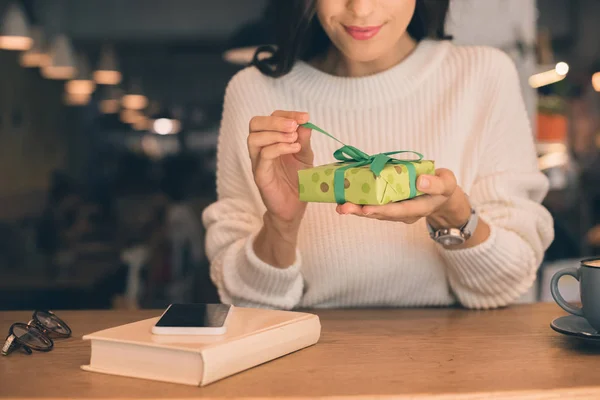 Vista parcial de la mujer joven desatando caja de regalo en la mesa con teléfono inteligente y taza de café en la cafetería - foto de stock