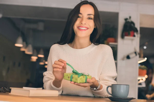 Alegre joven mujer desatando caja de regalo en la mesa con teléfono inteligente y taza de café en la cafetería - foto de stock