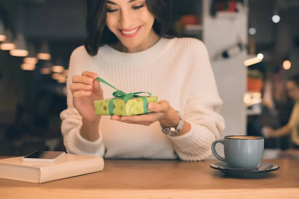 Sonriente mujer joven desatando caja de regalo en la mesa con teléfono inteligente y taza de café en la cafetería - foto de stock