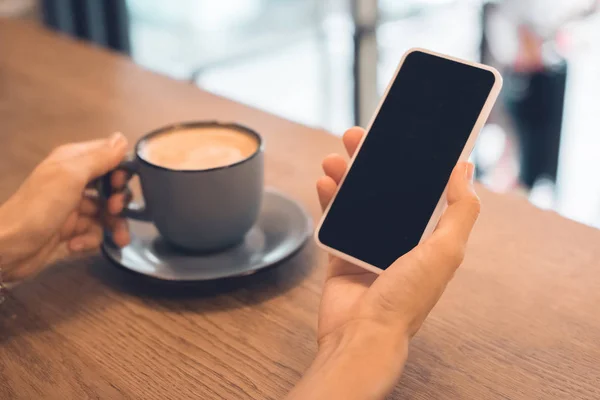 Обрезанный образ женщины с чашкой кофе с помощью смартфона с пустым экраном за столом в кафе — стоковое фото
