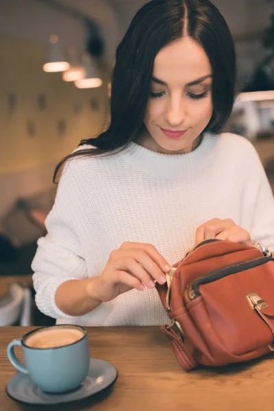 Atractiva mujer sacando dinero en efectivo de la bolsa en la mesa con taza de café en la cafetería - foto de stock