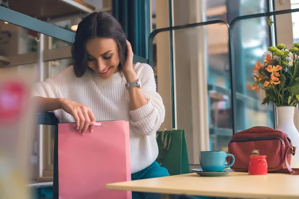 Foco seletivo de mulher sorridente olhando para o saco de compras à mesa com xícara de café no café — Fotografia de Stock
