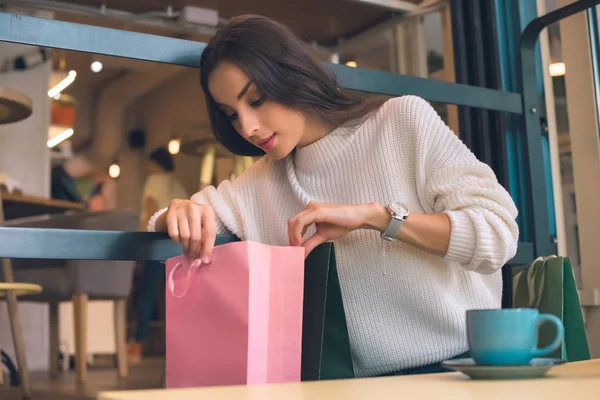 Mujer joven enfocada mirando en bolsa de compras en la mesa en la cafetería - foto de stock