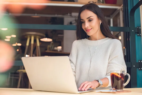 Female freelancer in earphones working on laptop at table with mulled wine in cafe — Stock Photo