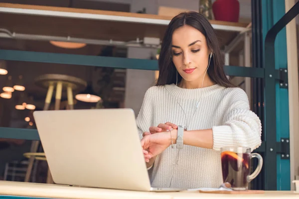 Young female freelancer in earphones checking wristwatch at table with laptop and mulled wine in cafe — Stock Photo