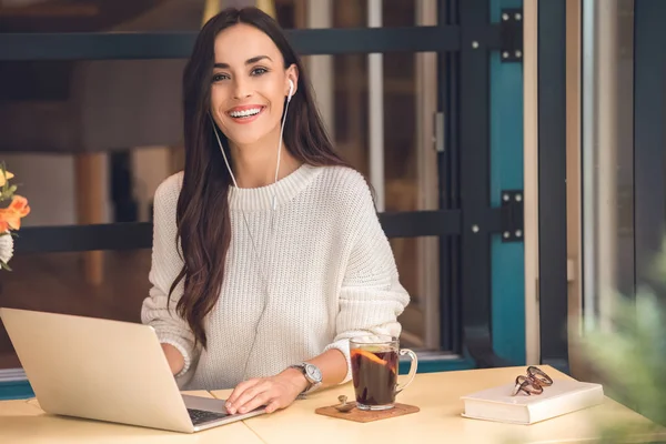 Attraktive junge Freiberuflerin mit Kopfhörern, die am Laptop arbeitet und bei Glühwein im Café in die Kamera schaut — Stockfoto