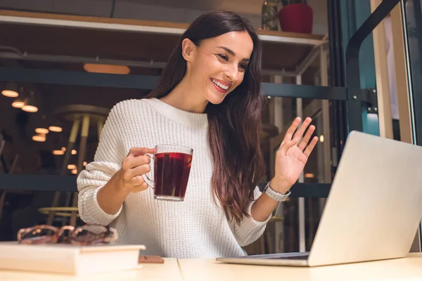 Щаслива молода жінка тримає чашку глінтвейну і махає вручну, маючи відеодзвінок на ноутбук за столом в кафе — стокове фото