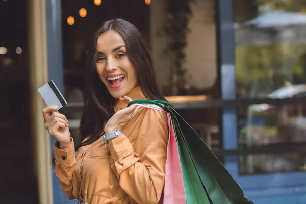 Hermosa mujer sonriente con bolsas de compras que muestran tarjeta de crédito en la calle de la ciudad - foto de stock