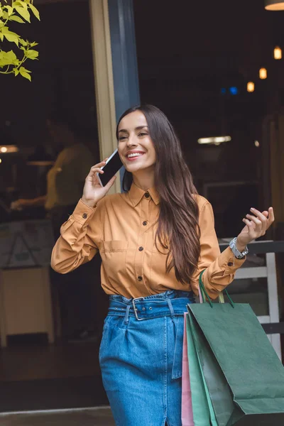 Mujer joven de moda con bolsas de compras hablando en el teléfono inteligente en la calle de la ciudad - foto de stock