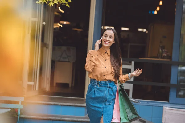 Selective focus of happy stylish young woman with shopping bags talking on smartphone at city street — Stock Photo
