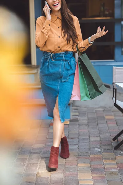 Vista parcial del elegante comprador femenino con bolsas de compras hablando en el teléfono inteligente en la calle de la ciudad - foto de stock