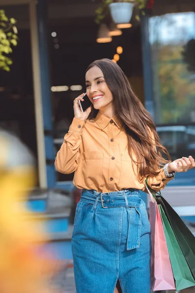 Feliz mujer joven con estilo con bolsas de compras hablando en el teléfono inteligente en la calle de la ciudad - foto de stock