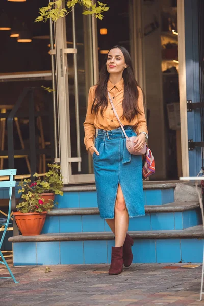 Attractive fashionable young woman with stylish handbag walking out from cafe at urban street — Stock Photo