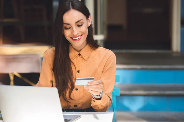 Sorrindo mulher elegante com cartão de crédito fazendo compras on-line no laptop à mesa no café na rua da cidade — Fotografia de Stock