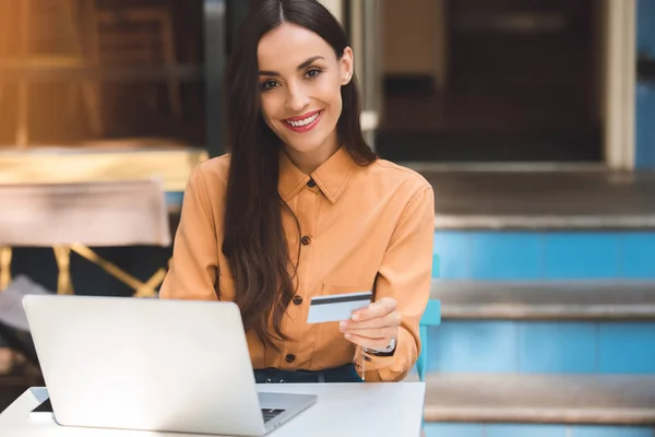 Attractive stylish woman with credit card doing online shopping on laptop at table in cafe at city street — Stock Photo