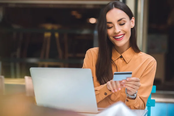 Selective focus of happy stylish woman with credit card doing online shopping on laptop at table in cafe at city street — Stock Photo