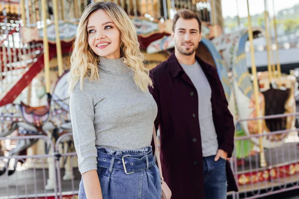 Smiling affectionate couple in autumn outfit looking away near carousel in amusement park — Stock Photo