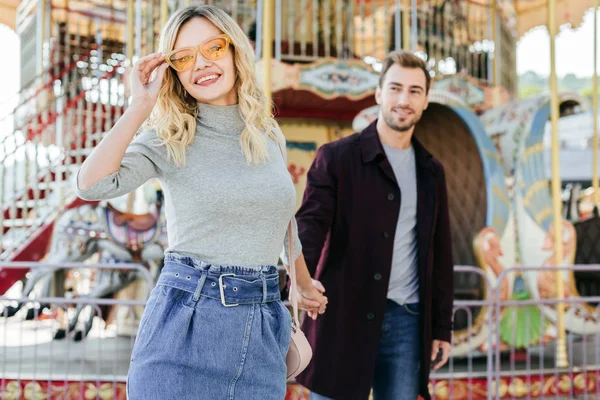 Sourire couple affectueux en tenue d'automne tenant la main près du carrousel dans le parc d'attractions, petite amie regardant la caméra — Photo de stock