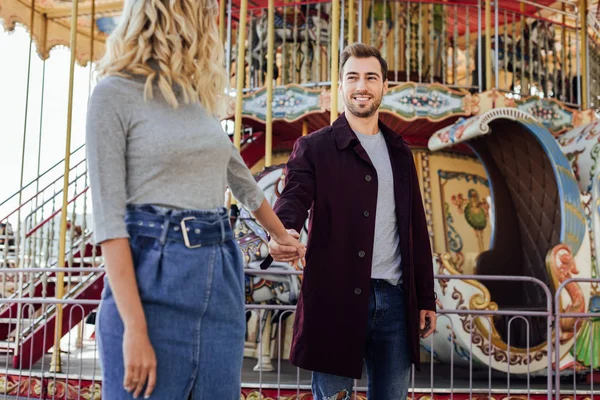 Affectionate couple in autumn outfit holding hands near carousel in amusement park and looking at each other — Stock Photo