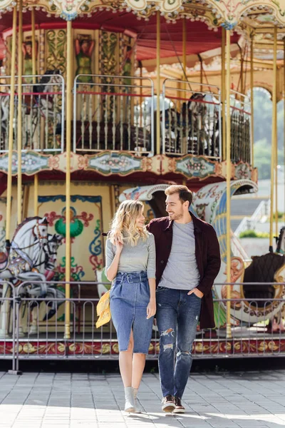 Ніжна пара в осінньому вбранні, що йде біля каруселі в парку розваг і дивиться один на одного — стокове фото