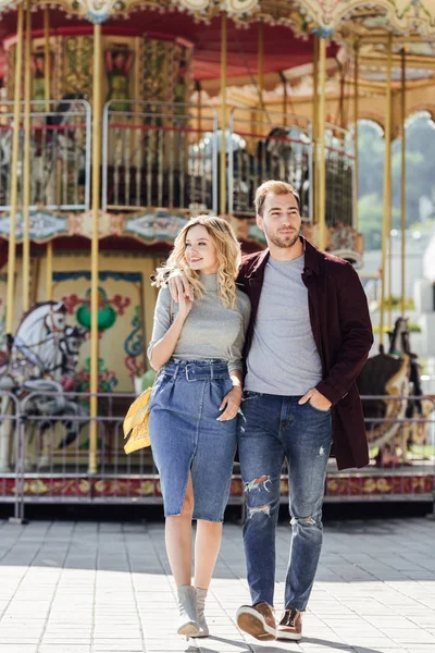 Pareja afectuosa en traje de otoño caminando y abrazándose cerca del carrusel en el parque de atracciones - foto de stock