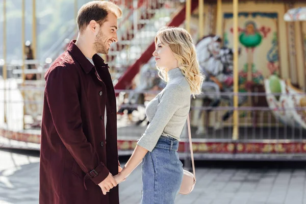 Affectionate couple in autumn outfit holding hands and looking at each other near carousel in amusement park — Stock Photo