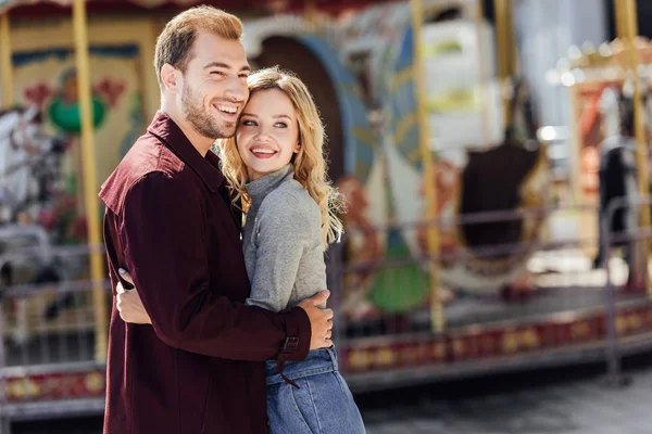 Sonriente pareja cariñosa en traje de otoño abrazándose cerca del carrusel en el parque de atracciones y mirando hacia otro lado - foto de stock